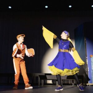 Two students performing on stage in costume during a middle school theater production under stage lighting.